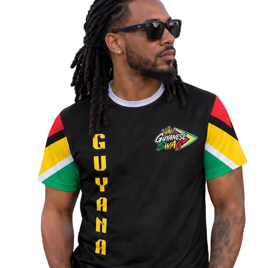 A man with dreadlocks and sunglasses wearing a black Guyana-themed t-shirt with red, yellow, and green sleeve details and 'GUYANA' printed vertically on the front, standing in a crowded outdoor street setting.