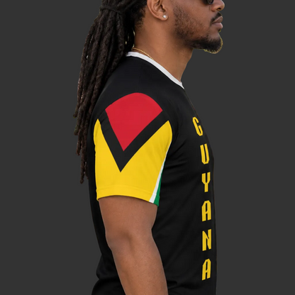 A man wearing a black Guyana-themed t-shirt with red, yellow, and black graphic details on the sleeve and yellow 'GUYANA' text down the side, standing outdoors in a crowd.